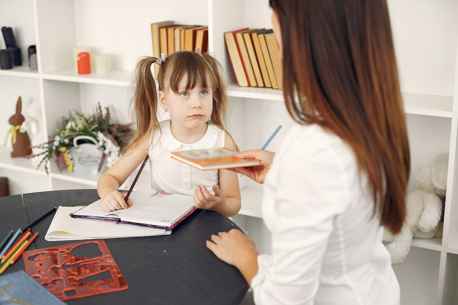Niña pequeña sentada a la mesa escribiendo en un cuaderno con lápiz mientras toma un libro de texto de una joven maestra particular vestida con una camisa blanca durante la educación en casa, apoyada en un estante con libros de texto.